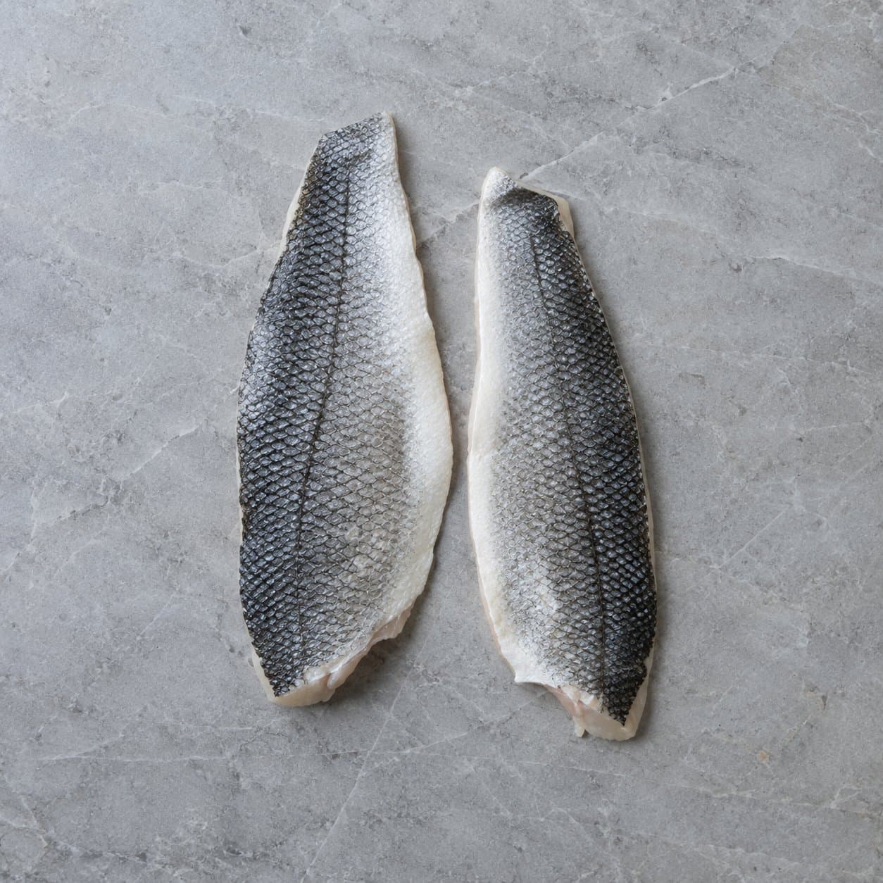 Two raw sea bass fillets, skin side up, on grey stone